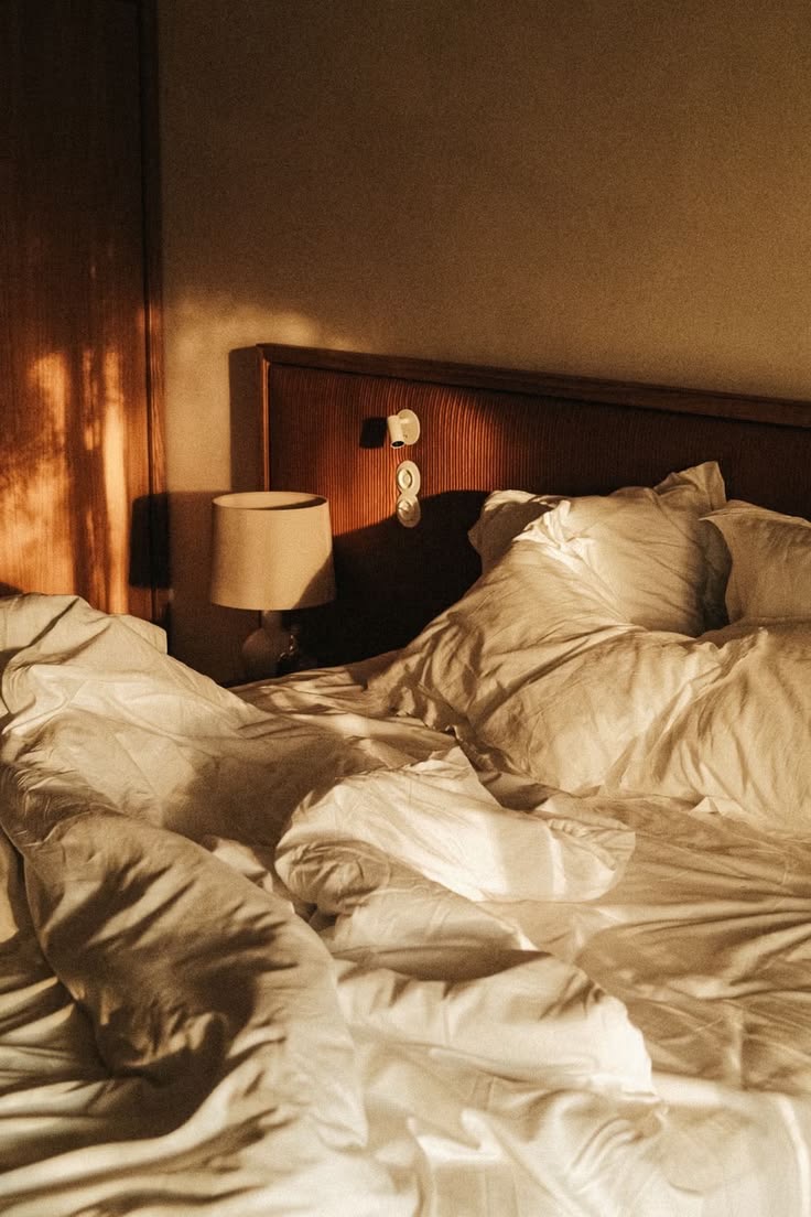 Lit défait aux draps de lin clair, tête de lit de bois cannelé et lampe de chevet, dans la lumière du matin